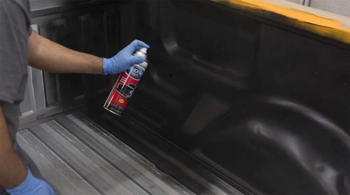 Person in blue gloves spraying protective coating inside a black truck bed with a red aerosol product