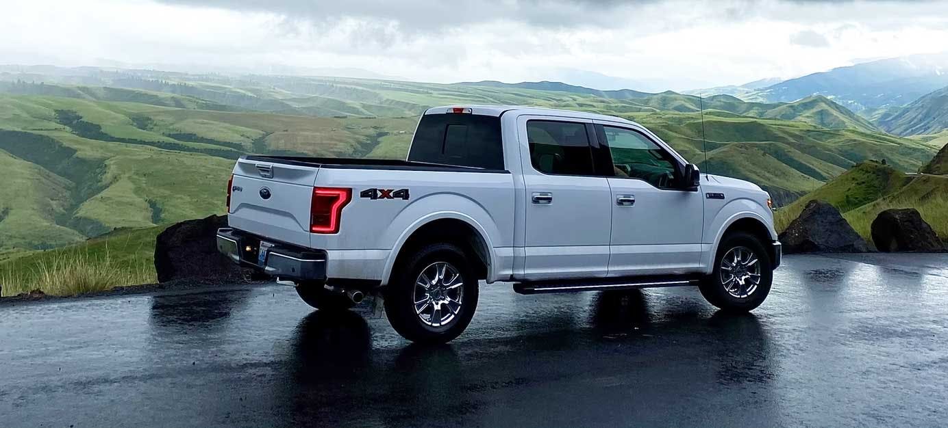 White Ford F-150 4x4 crew cab parked on wet pavement with rolling green mountains and grassland in background