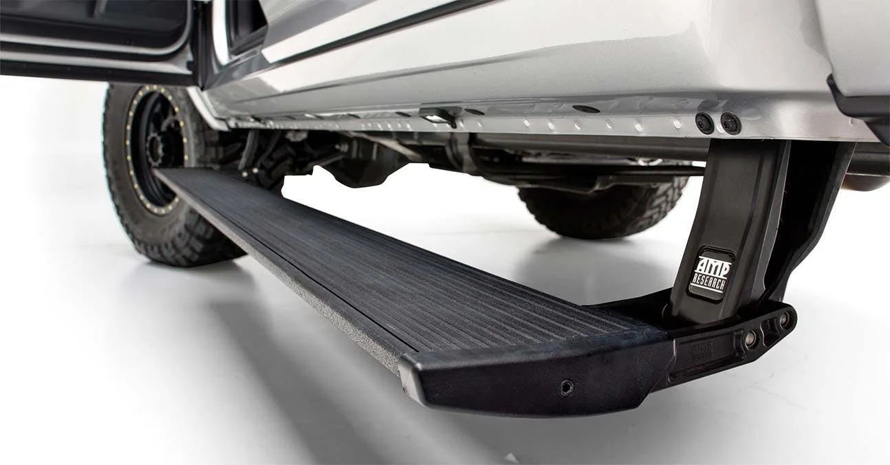 Black AMP Research PowerStep running board extended from the side of a white truck, with textured tread surface and