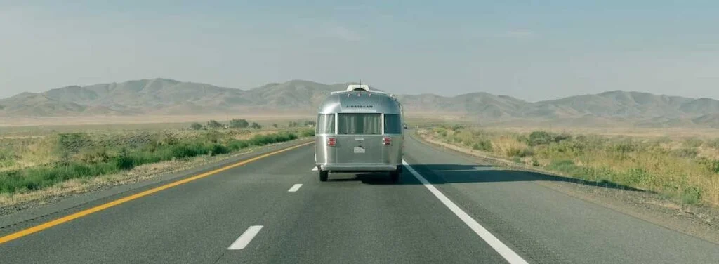 Silver Airstream travel trailer being towed on a straight desert highway with mountains in the distance