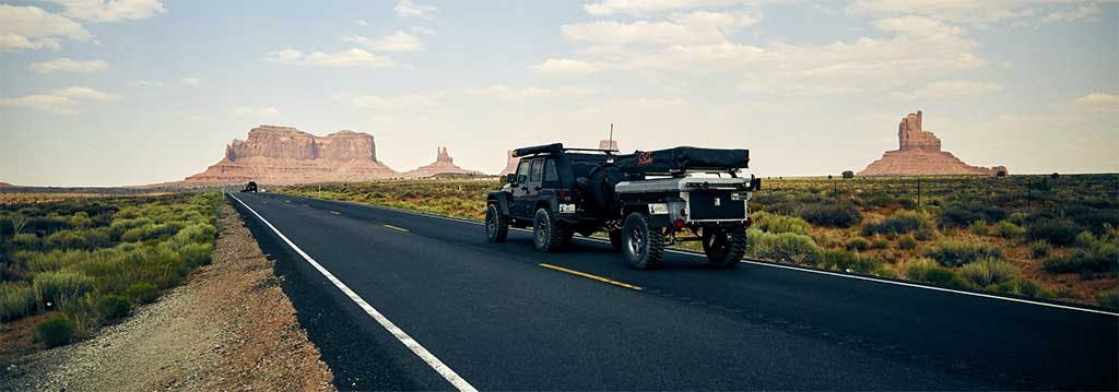 Black pickup truck towing a white utility trailer on a desert highway with buttes in the background