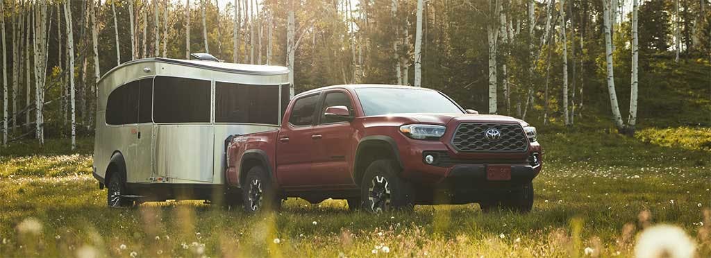Red Toyota Tacoma pickup truck towing a silver travel trailer through a wildflower meadow with birch forest backdrop