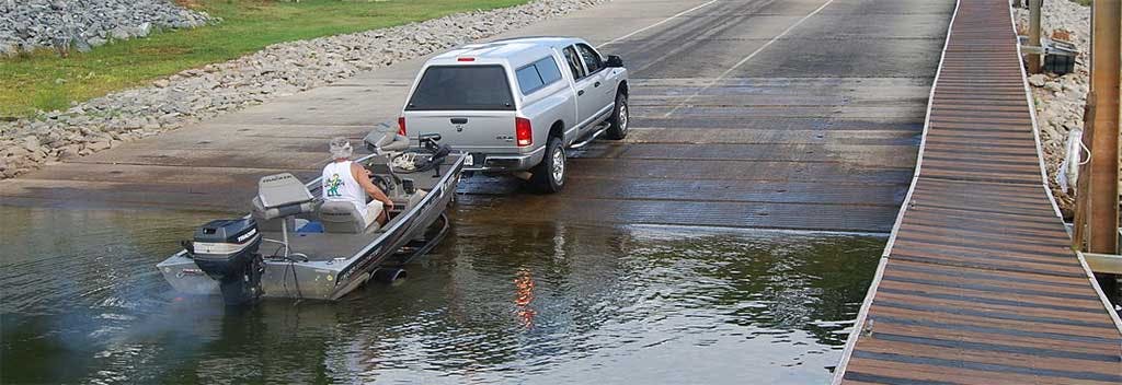 White pickup truck parked on boat ramp with fishing boat in water and wooden dock extending to the right.