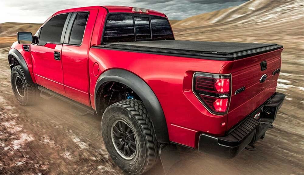 Red pickup truck with black tonneau cover on bed, parked on rocky terrain with desert landscape behind it.