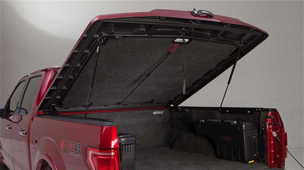 Red truck bed with black hard folding tonneau cover open, revealing dark carpet interior and metal support struts