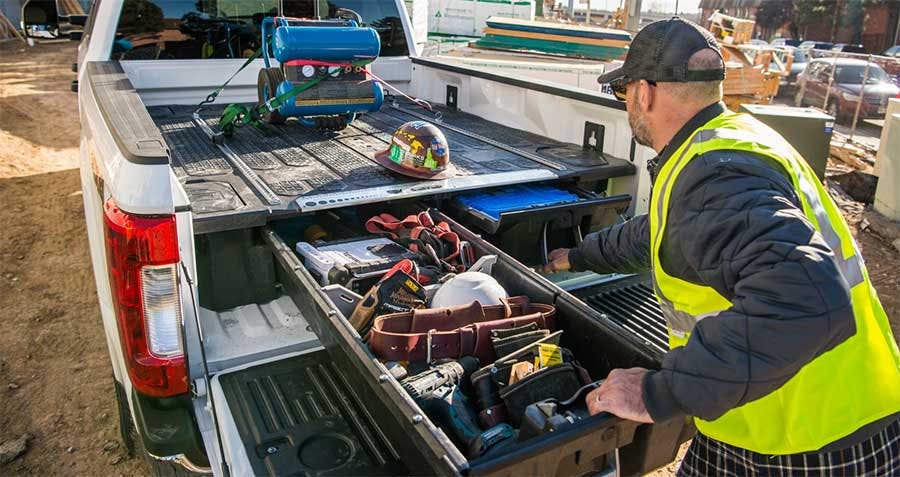 Worker in yellow safety vest accessing sliding drawers installed in white pickup truck bed loaded with tools and equipment