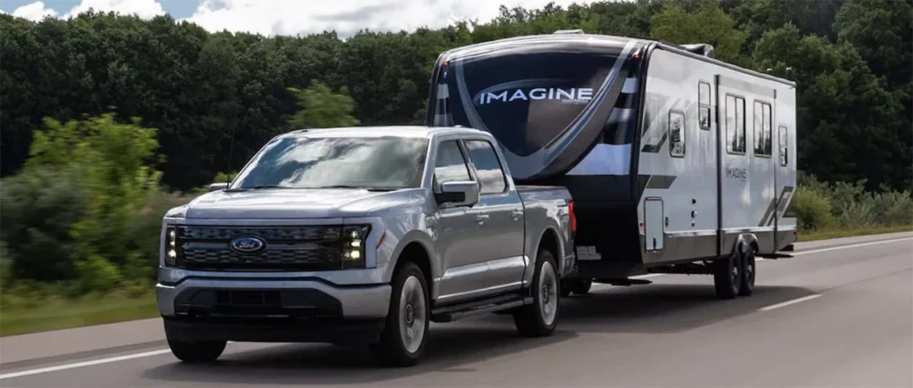 Silver Ford F-150 towing a white and black Imagine travel trailer on a highway with trees in the background.