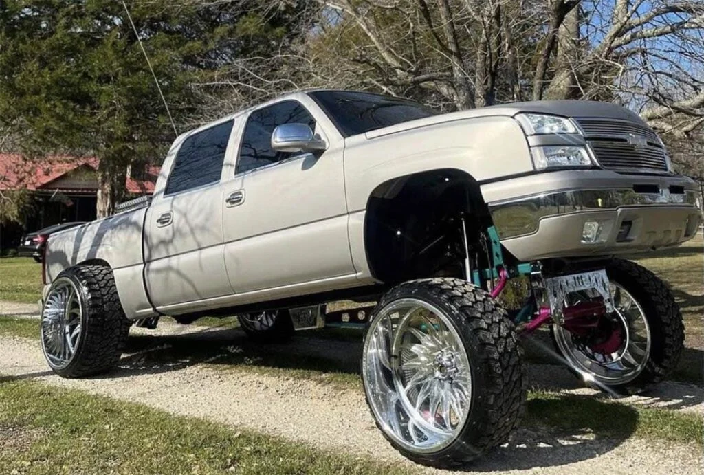 White lifted truck with exaggerated front-end lift and lowered rear parked on gravel driveway with bare trees behind
