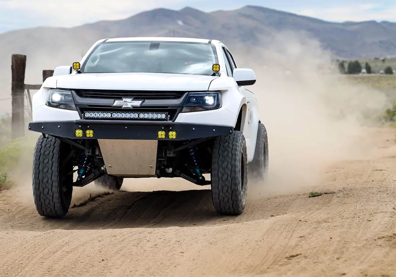 White Chevrolet truck with extreme front lift kit driving on dirt road, kicking up dust, equipped with LED light bar and