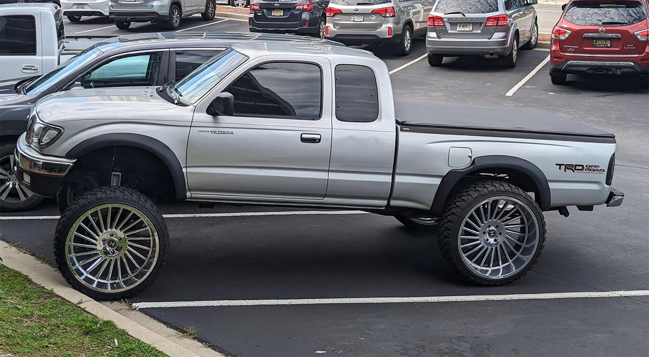 Silver Toyota Tacoma on tall aftermarket wheels parked in lot, rear end lowered with front suspension raised higher.