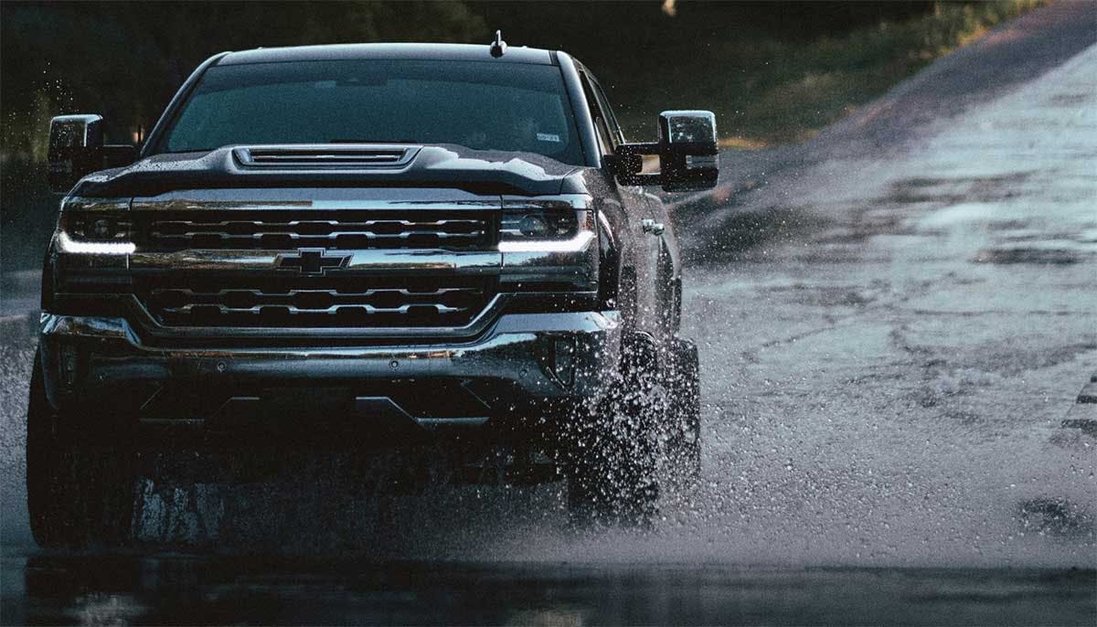 Black Chevy truck splashing through muddy water on an unpaved road with another vehicle trailing behind.