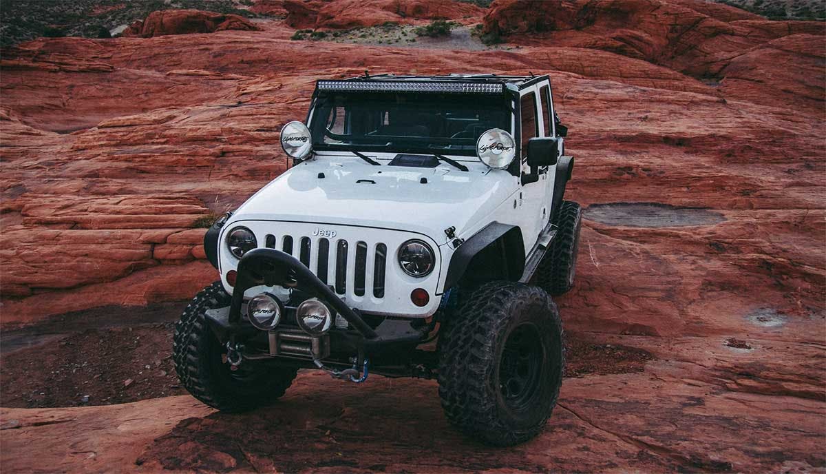 White Jeep with lift kit, oversized black tires, and roof-mounted light bar parked on red rock terrain.