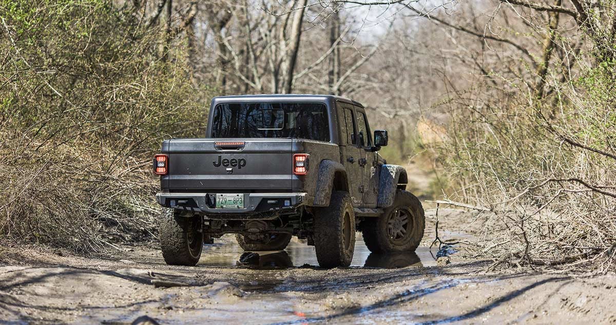 Jeep Wrangler with lifted suspension driving through muddy water on a wooded trail, showing modified off-road setup